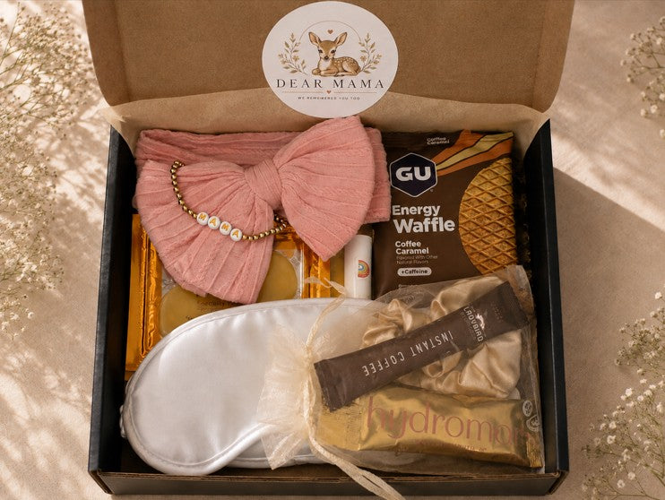 A curated gift box labeled 'Dear Mama' opened to reveal a pink pleated bow headband with pearl beads, a GU Energy Waffle coffee bar, a white satin sleep mask, and a gold-toned bath towel wrapped in cellophane. The box is photographed from above on a beige fabric surface with soft sunlight and leaf shadows.