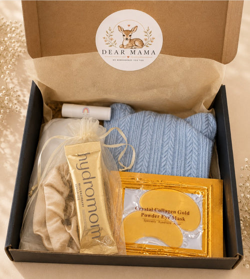 A curated gift box set from Dear Mama, featuring a light blue knit sweater, a gold tube of HydroMom moisturizer, a gold packet of Crystal Collagen Gold Powder Eye Mask, and a beige tassel hair tie, all arranged inside a black box with a brown cardboard insert and a circular deer logo sticker on the inside lid.